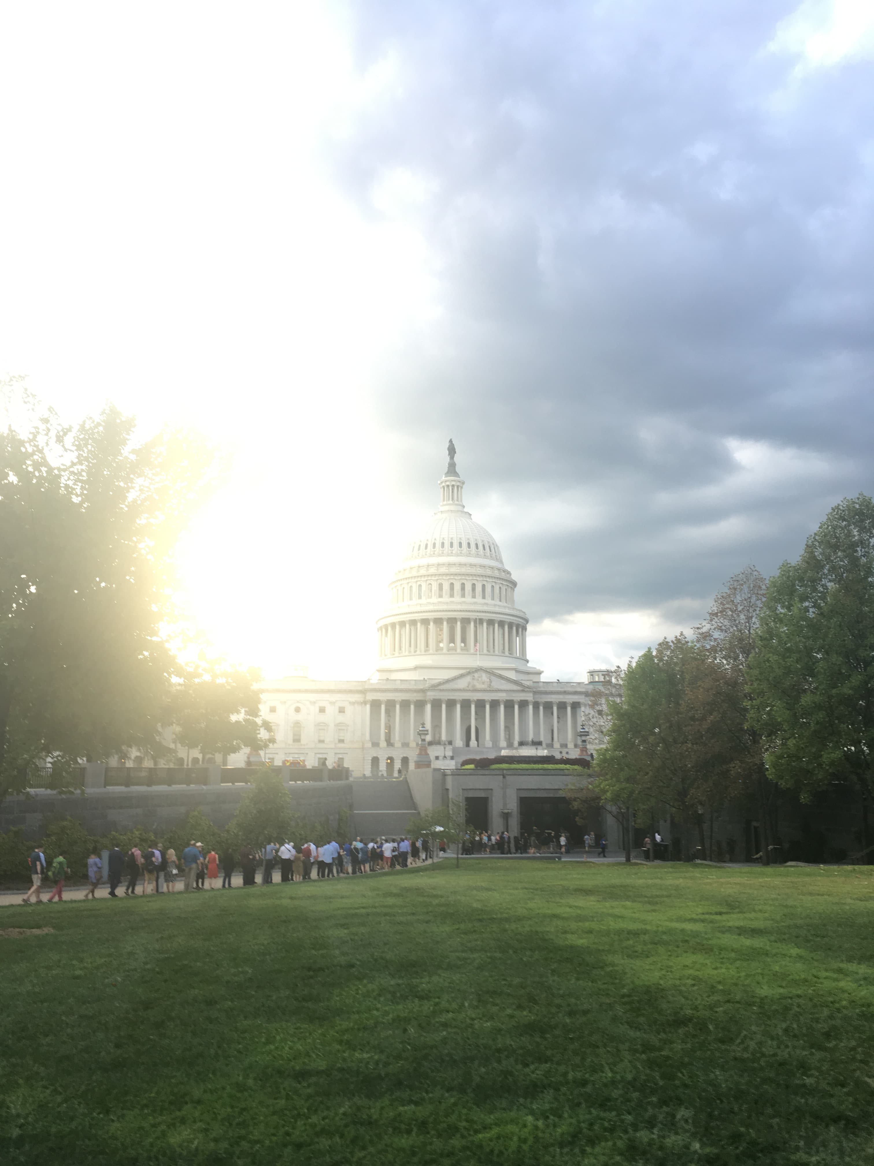 U.S. Capitol dome under dramatic clouds with bright sunlight flare and a green lawn.